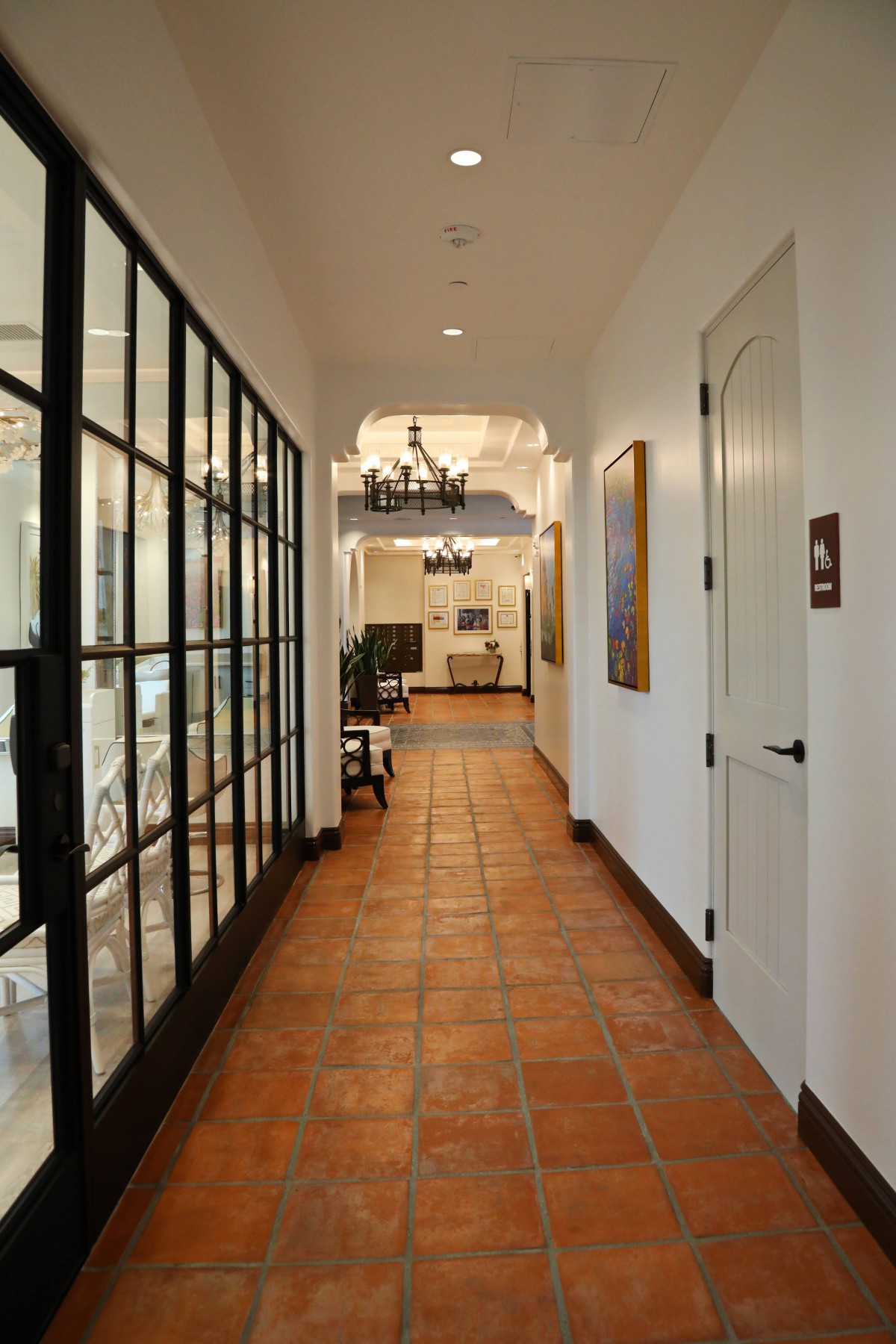A well-lit hallway with terracotta tiles, glass panel doors, and chandeliers.