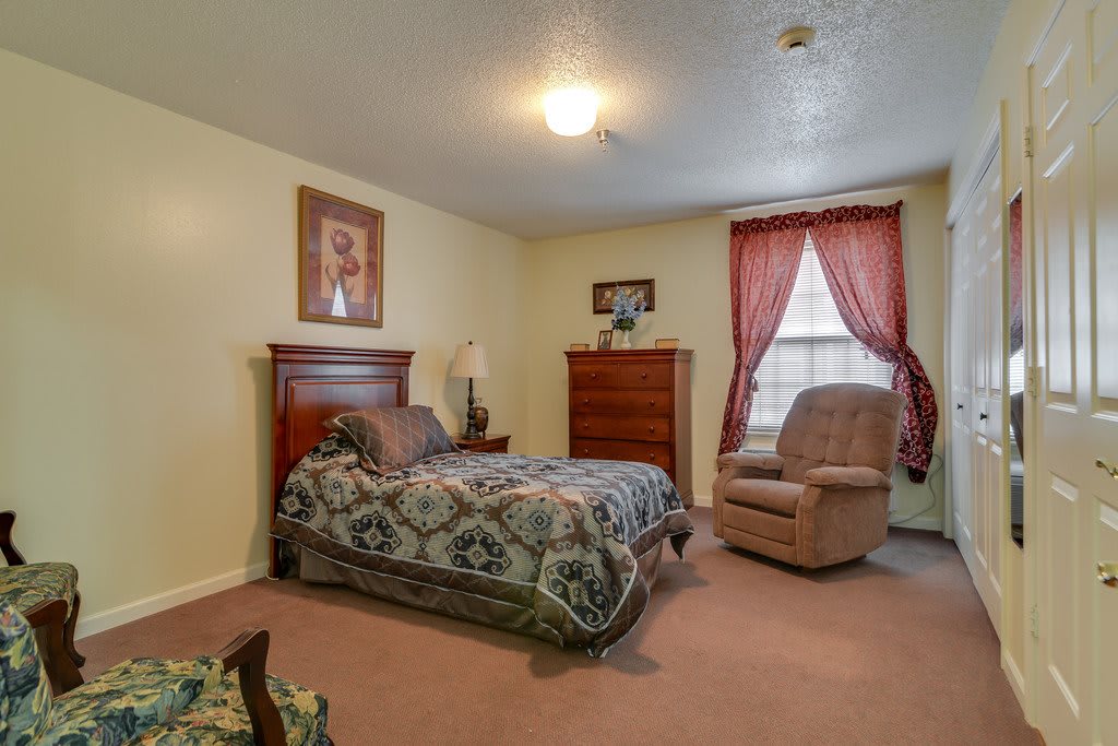 A cozy bedroom with a bed, dresser, and armchair near a window with red curtains.