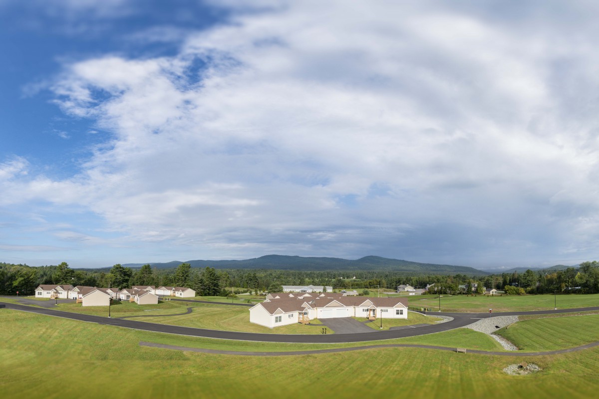 A scenic view of a suburban neighborhood with houses, wide open grass fields, and mountains in the background under a cloudy sky.