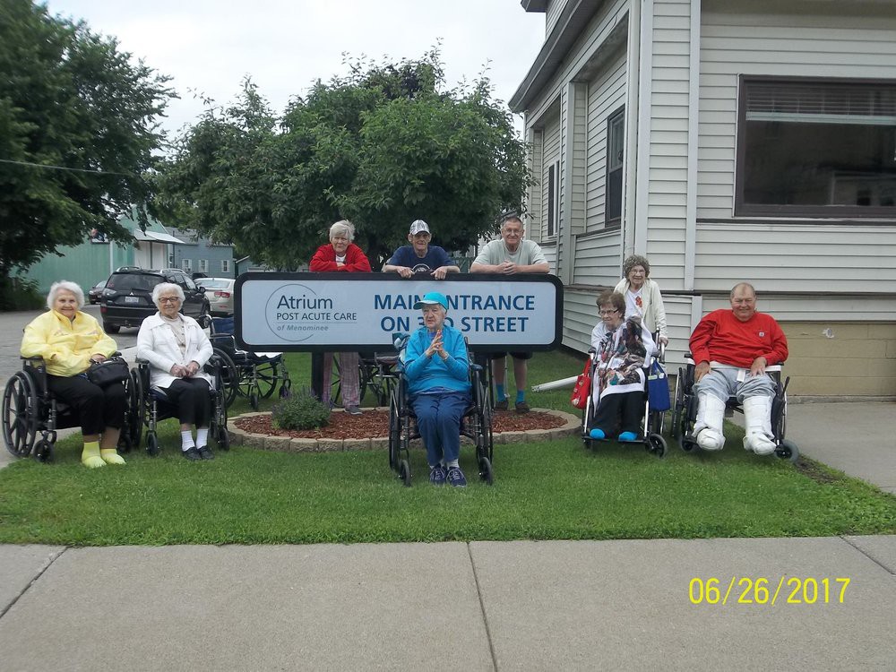 A group of elderly individuals sitting outside near a building with a sign, some in wheelchairs.
