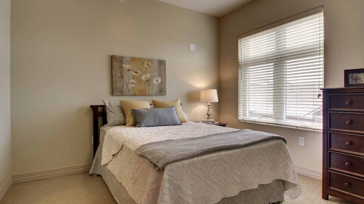 A cozy bedroom with a neatly made bed, side lamp, and large window providing natural light.