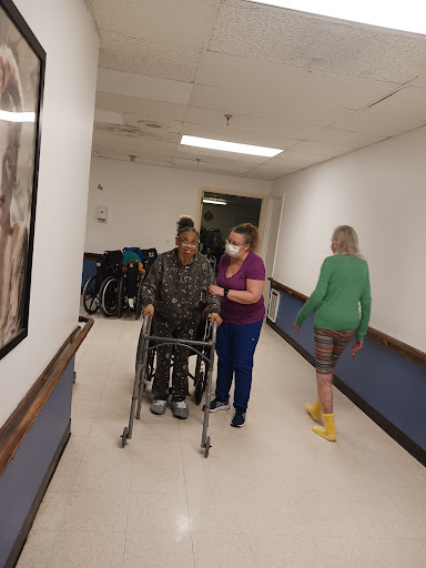 A caregiver assists a patient in a wheelchair while another caregiver walks nearby in a hospital hallway.