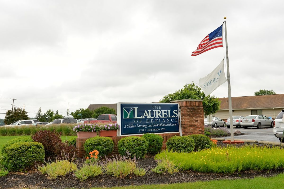 The entrance sign of The Laurels of Defiance nursing home with an American flag waving under a cloudy sky.
