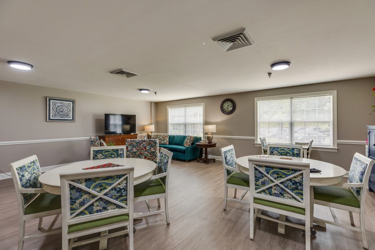 A cozy living room with multiple tables and chairs, a television, and large windows providing natural light.