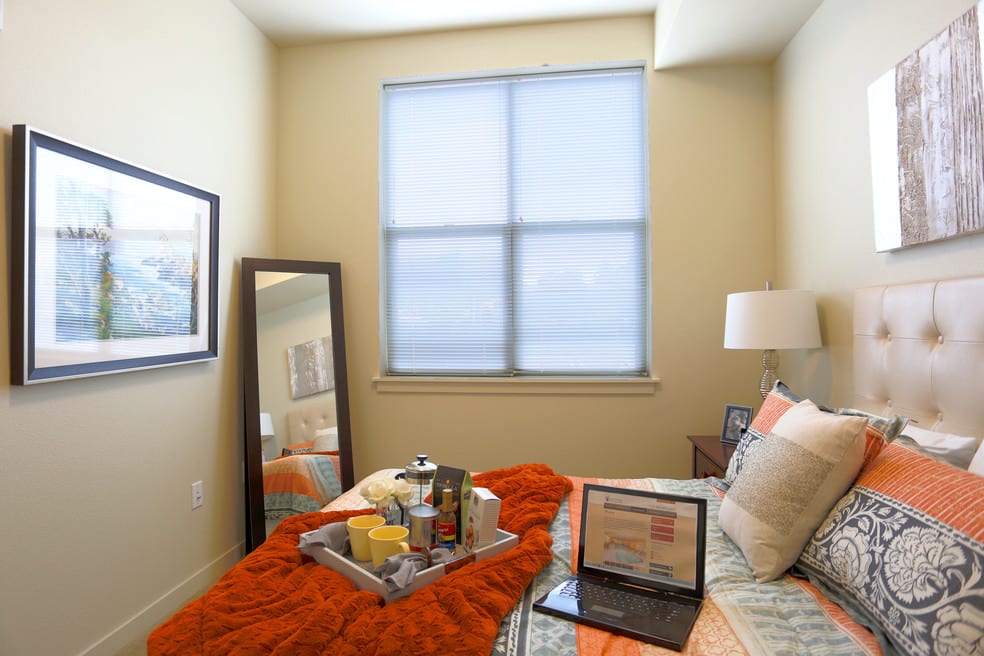 A cozy bedroom with a neatly arranged bed, featuring a laptop, mirror, and window.
