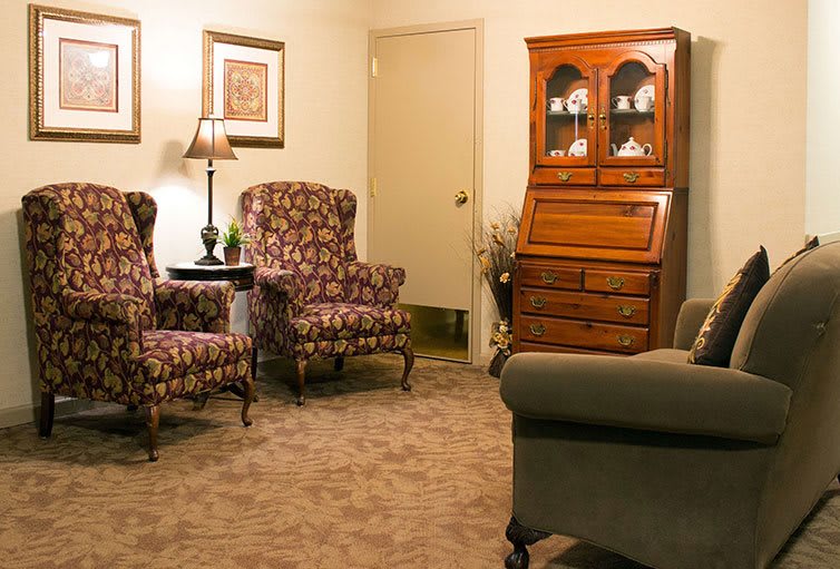 A cozy living room with patterned armchairs, a wooden cabinet, and a lamp.