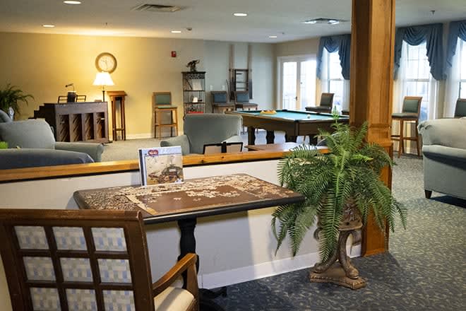 A cozy lounge area with a table set for a puzzle, surrounded by comfortable seating and a pool table in the background.