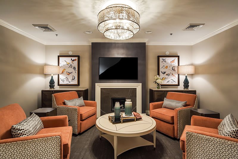A modern living room with orange chairs, a central coffee table, and a chandelier above, features a fireplace and television.