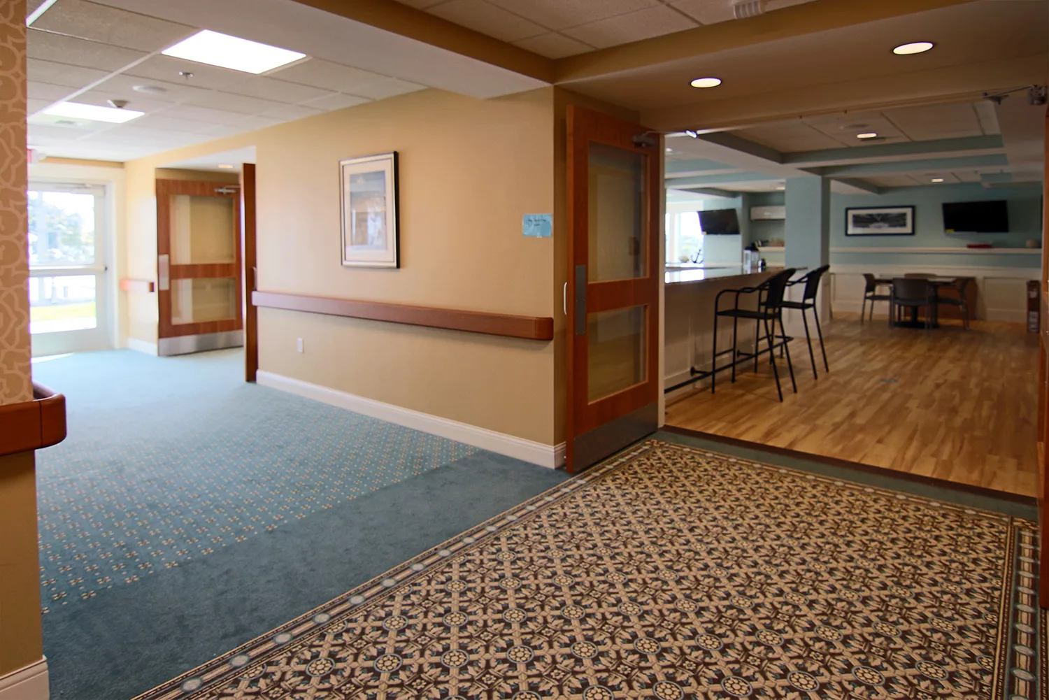 A hallway with carpeted flooring leading to a kitchen area with bar stools.
