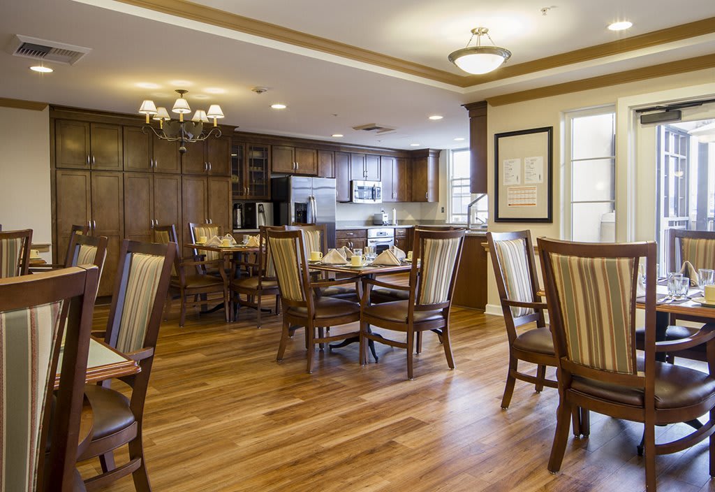 A cozy dining room with wooden flooring, neatly arranged tables and chairs, and a kitchen area in the background.