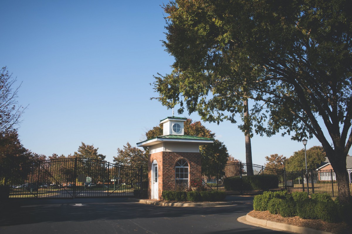 A quaint brick guardhouse situated amidst a tree-lined driveway under a clear blue sky.