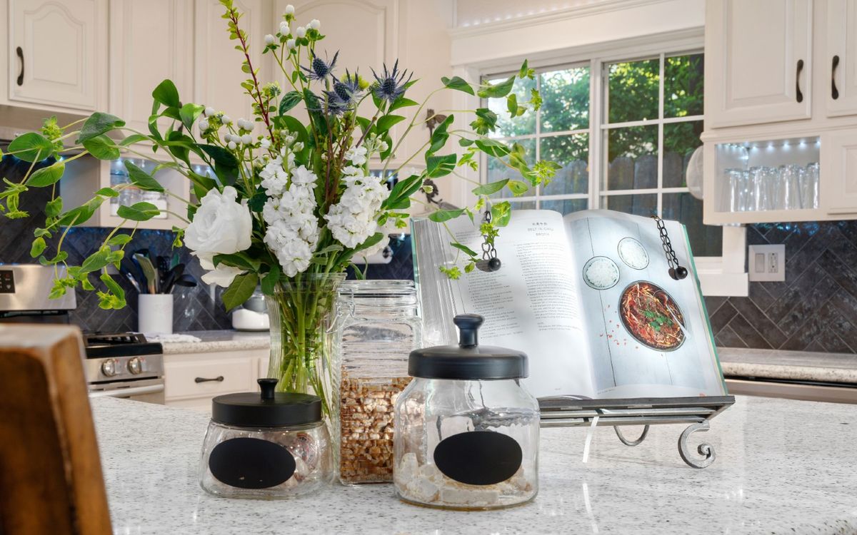 A bright kitchen countertop featuring a vase of flowers, storage jars, and an open cookbook.