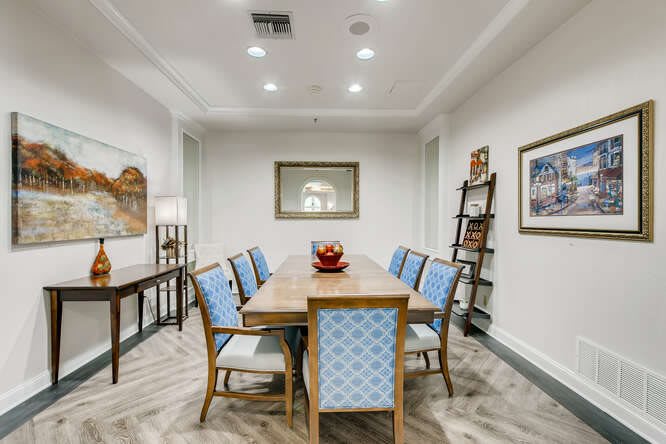 A modern dining room with a wooden table, blue upholstered chairs, artwork on the walls, and a bookshelf in the corner.
