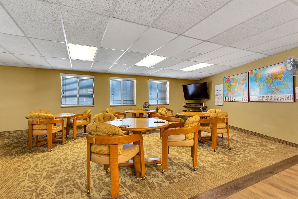 A well-lit meeting room with multiple tables and chairs, featuring a wall-mounted television and world maps.