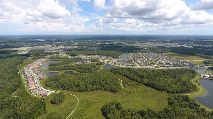 An aerial view of a suburban neighborhood surrounded by forests and lakes under a partly cloudy sky.
