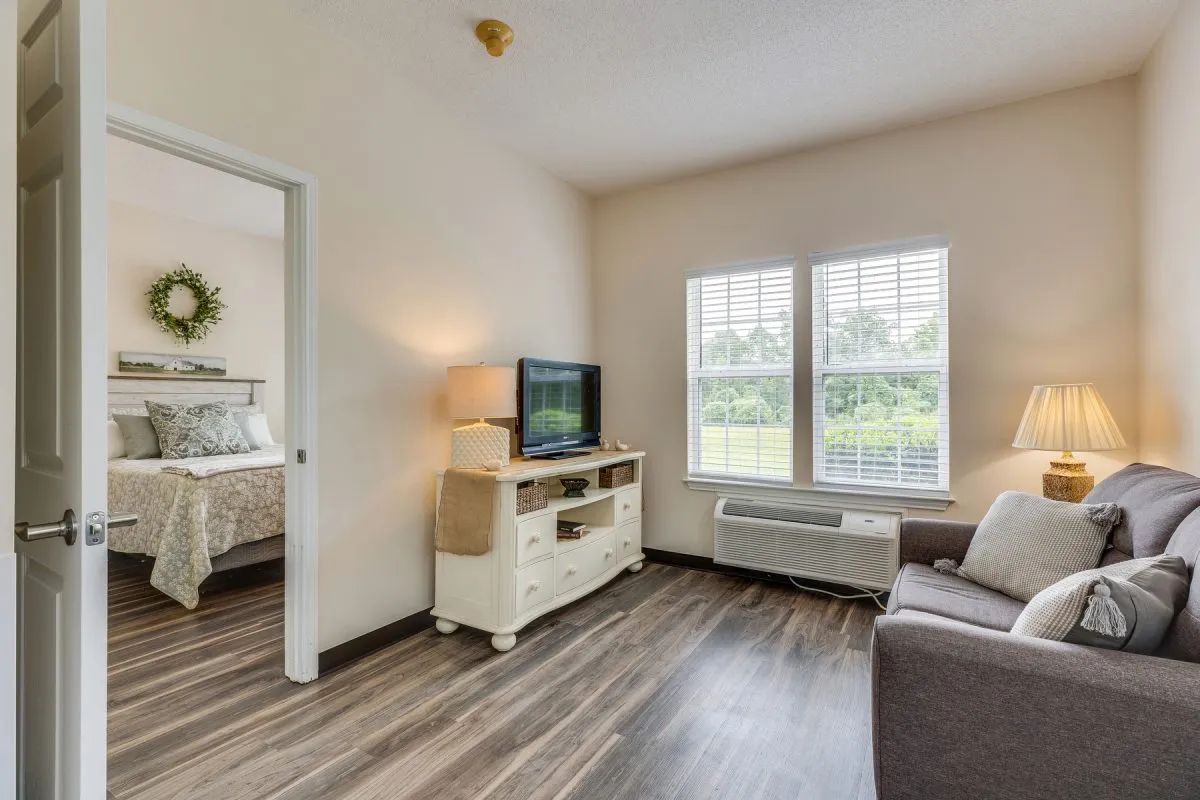 A cozy living room with a sofa, television, and large windows, featuring wooden flooring.