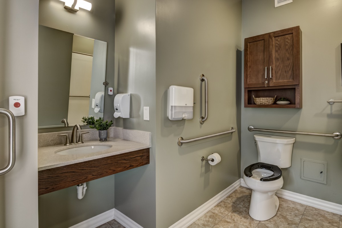 A modern bathroom with a sink, toilet, and storage cabinets, featuring accessibility handrails and a wall-mounted towel dispenser.