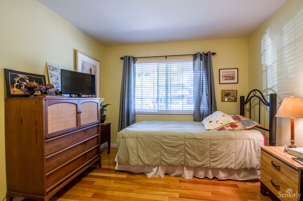 A cozy bedroom with a bed, wooden dresser, and a television next to a window with curtains.