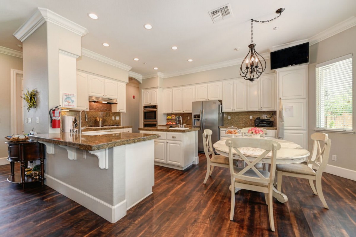 A modern kitchen with white cabinets and a dining area, featuring hardwood floors and elegant chandelier lighting.