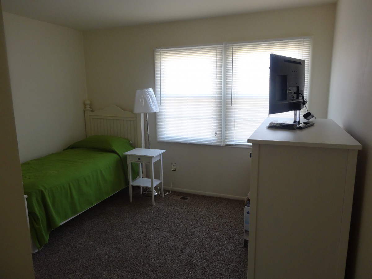 A minimalist bedroom features a single bed with green bedding, a small side table with a lamp, a dresser topped with a television, and natural light coming through the window.