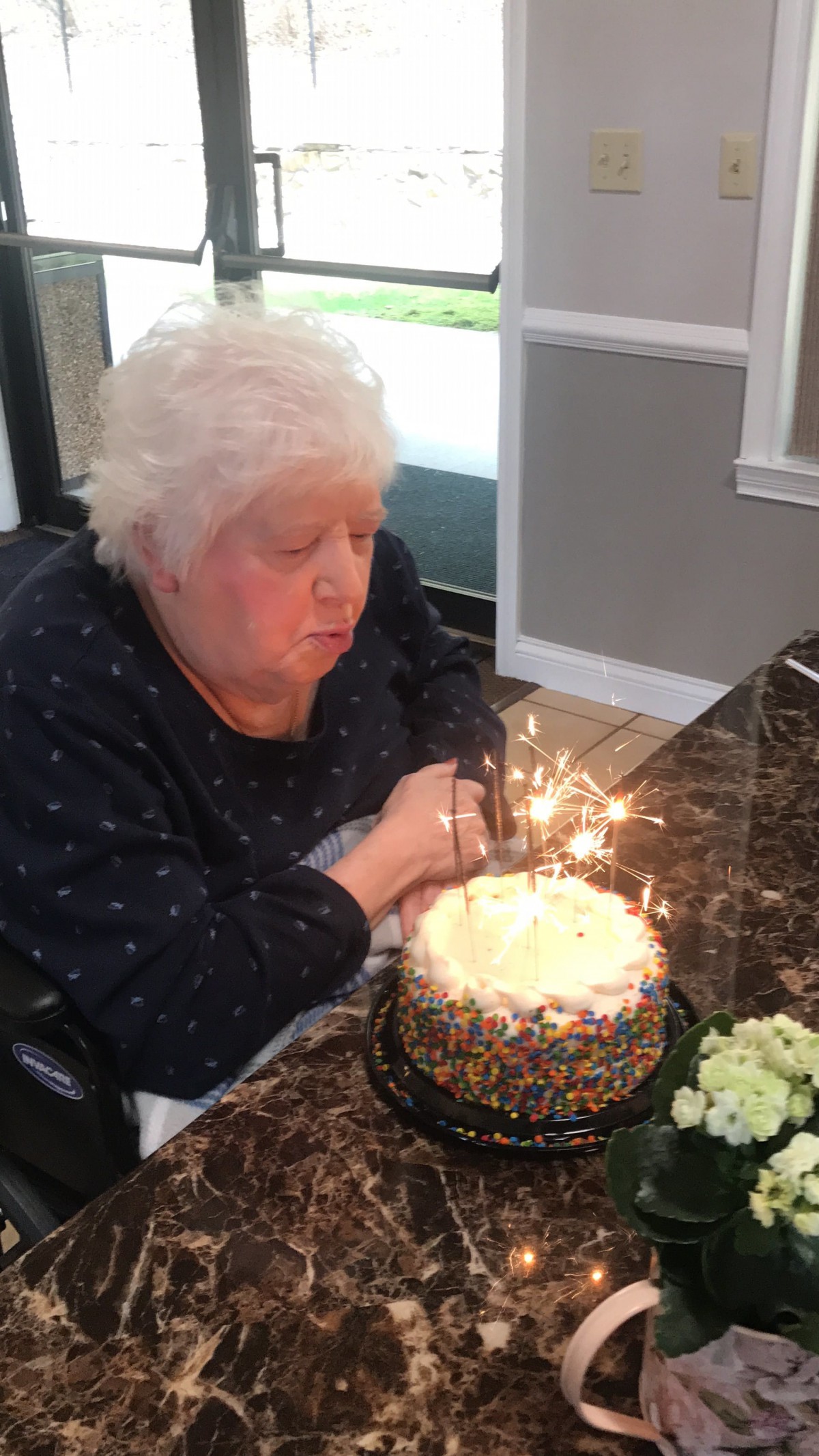 An elderly woman blowing out candles on a birthday cake with sparklers.