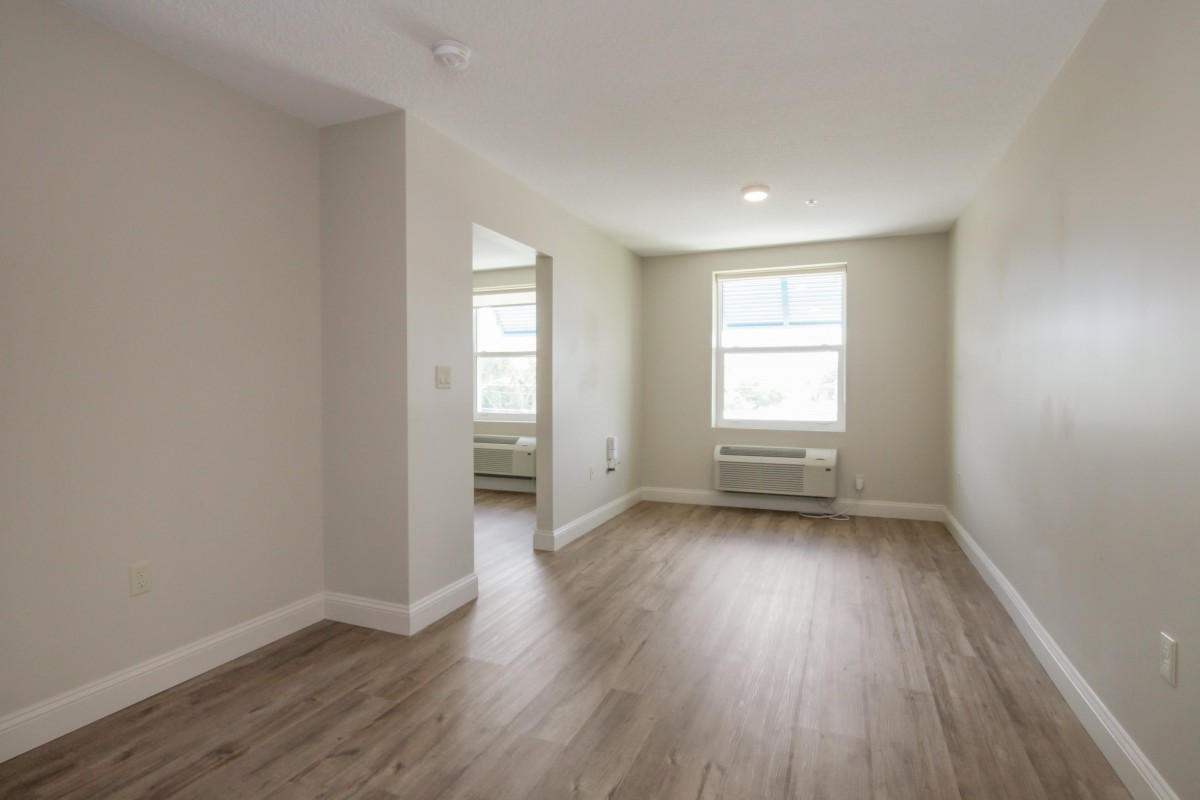 A bright, empty apartment room with windows and wooden flooring.