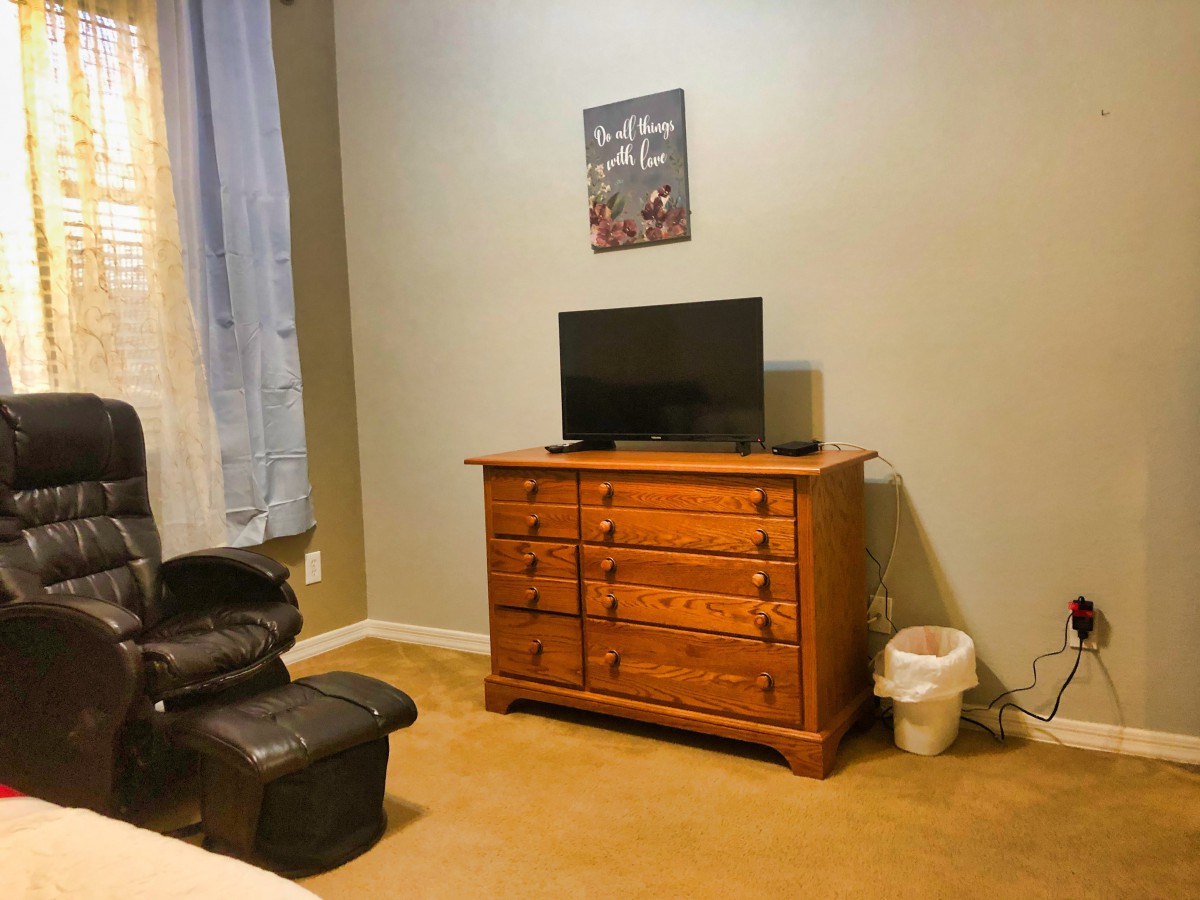A cozy bedroom corner features a wooden dresser with a TV on top, a comfortable chair, and wall art.