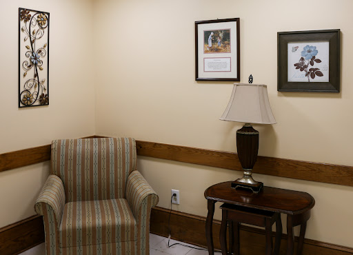 A cozy waiting area featuring a striped armchair, a side table with a lamp, and framed pictures on the walls.