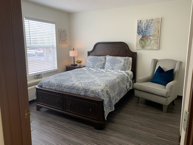 A cozy bedroom with a neatly made bed, a lamp on the bedside table, an armchair, and artwork on the wall.