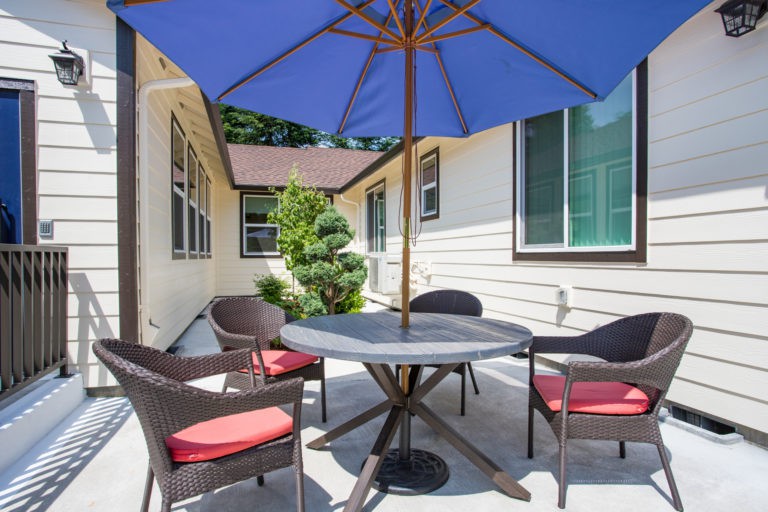A cozy patio setup with a table, chairs, and a large blue umbrella in a sunny outdoor space.