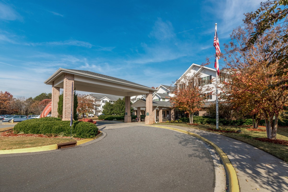 A well-maintained building entrance with a covered driveway, surrounded by landscaped gardens and a flagpole on a clear autumn day.