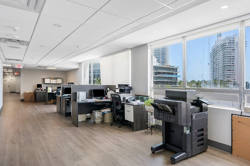 A modern office space featuring cubicles with computers and printers, illuminated by natural light coming through large windows.