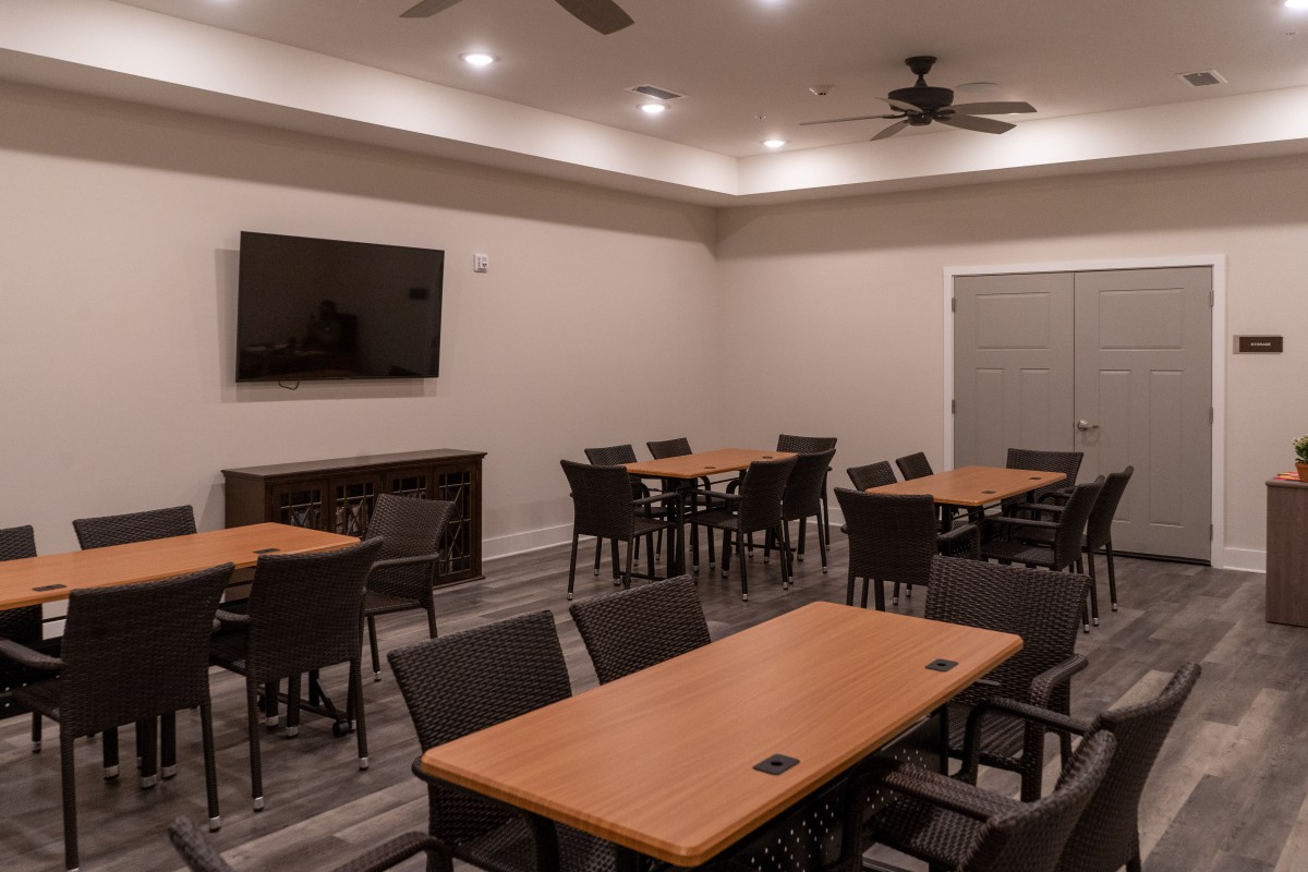 A well-lit meeting room with multiple tables and chairs, featuring a large television on the wall.