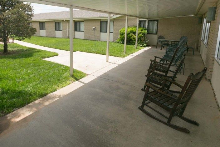 A covered patio with rocking chairs overlooking a well-maintained lawn in front of a building.