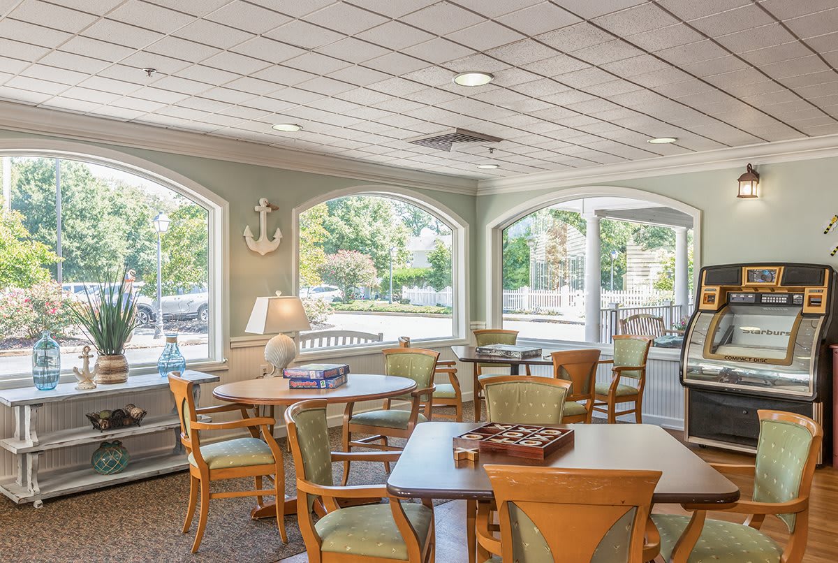 A bright community room with several tables and chairs, featuring large windows and nautical decor.