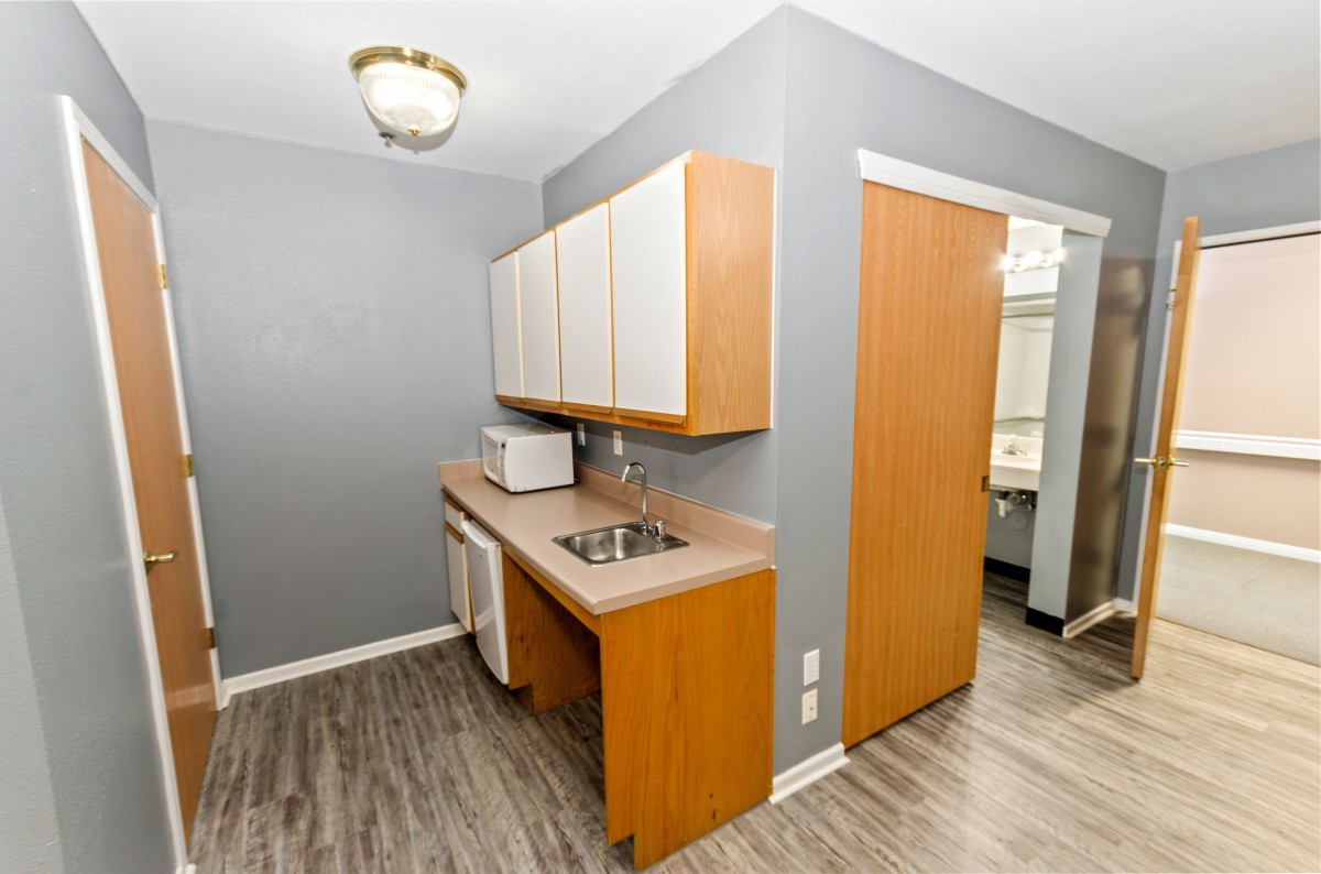 A small kitchen area with wooden cabinets and a sink, featuring a microwave on the countertop and a doorway leading to another room.
