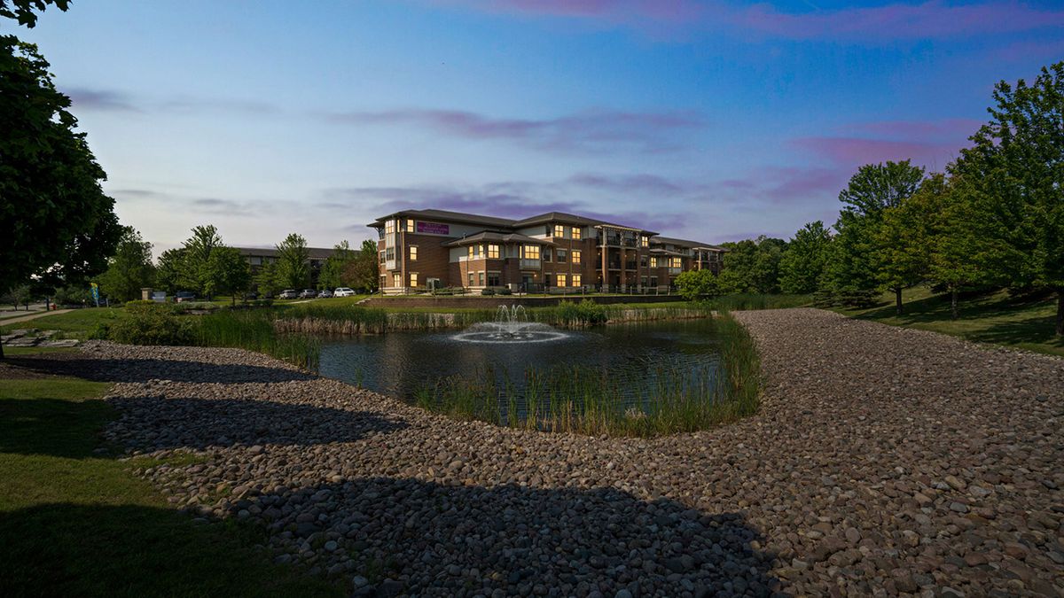 A scenic view of a residential building by a pond surrounded by greenery.