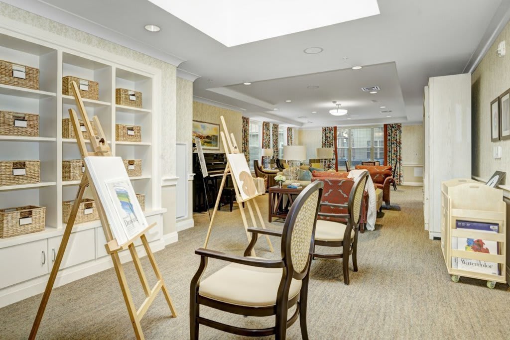 A well-lit interior space featuring an art gallery setup with chairs and easels displaying paintings.