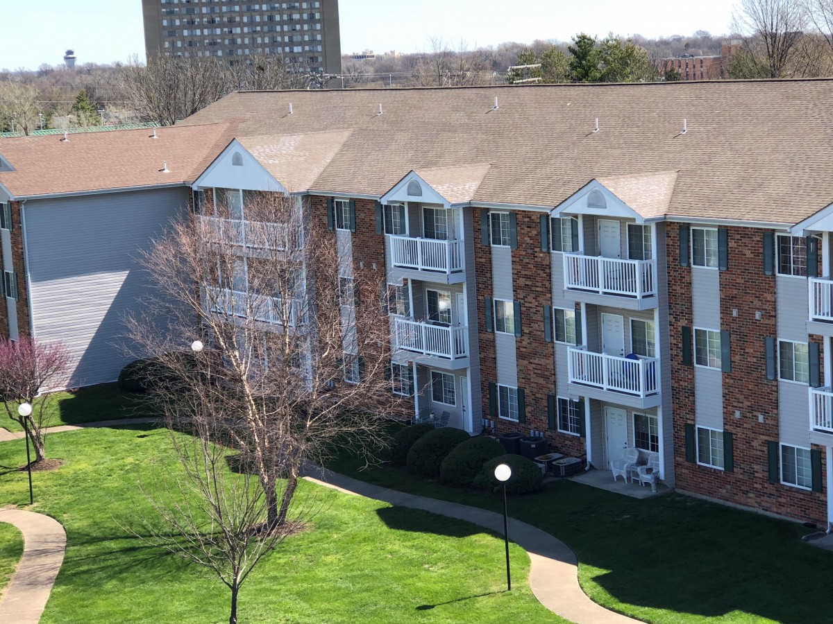 A multi-story apartment building with brick exterior and balconies, surrounded by green lawns and trees.