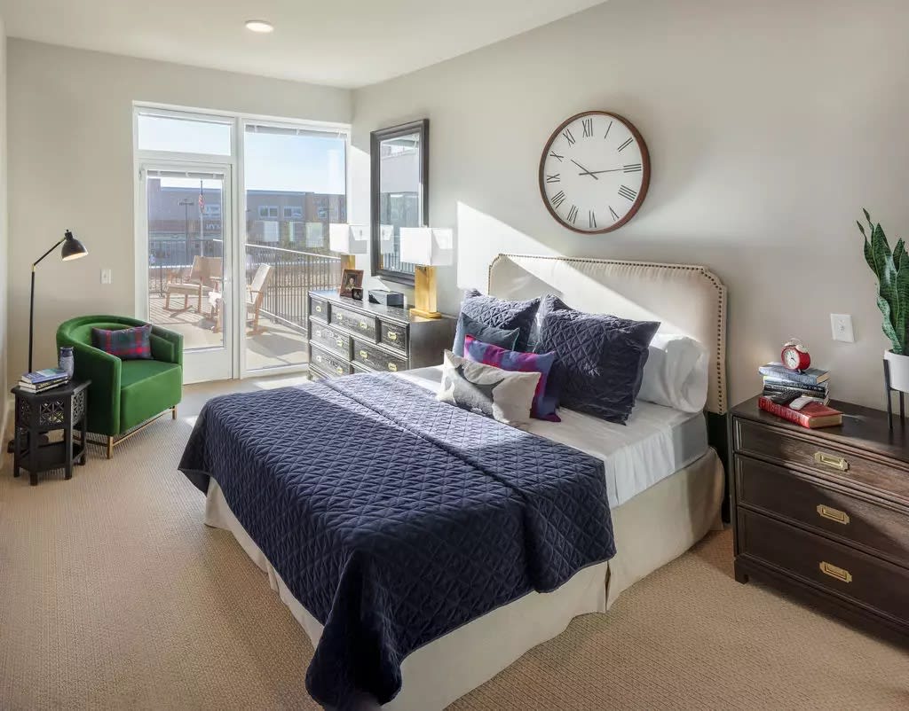 A modern bedroom with elegant decor, featuring a neatly made bed, large windows allowing natural light, and a clock on the wall.
