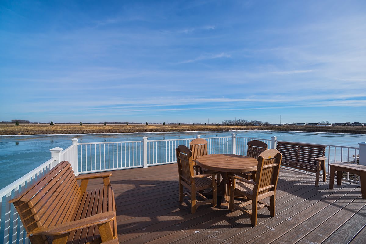 A scenic view of a waterfront deck with wooden chairs and table under a clear blue sky.
