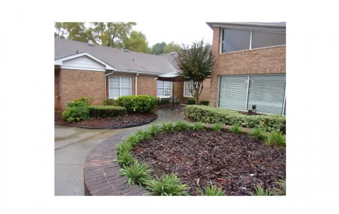 A residential brick house with a landscaped yard and shrubs along a concrete pathway.