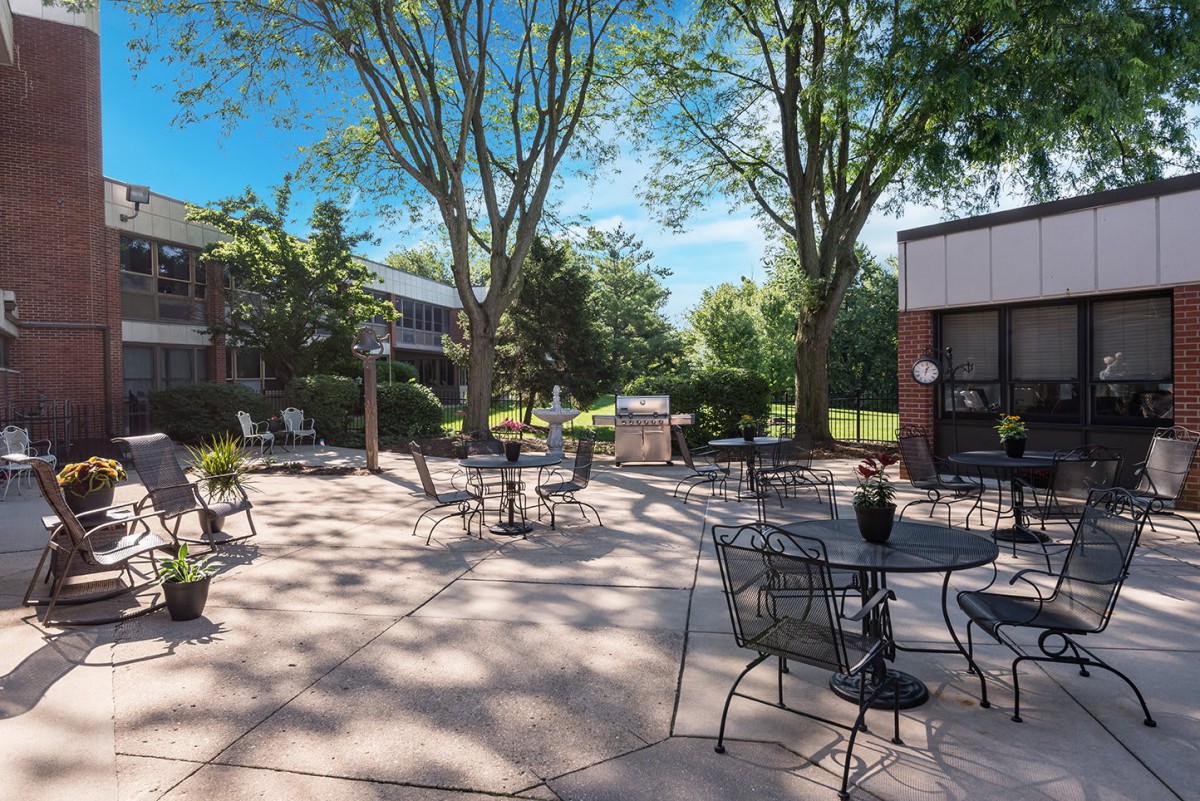 A serene outdoor patio area with tables and chairs, shaded by trees on a sunny day.