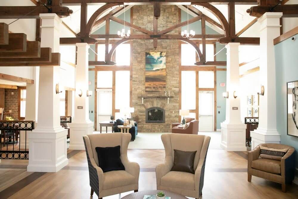 A cozy living room with two armchairs facing a stone fireplace, flanked by large windows and exposed wooden beams.
