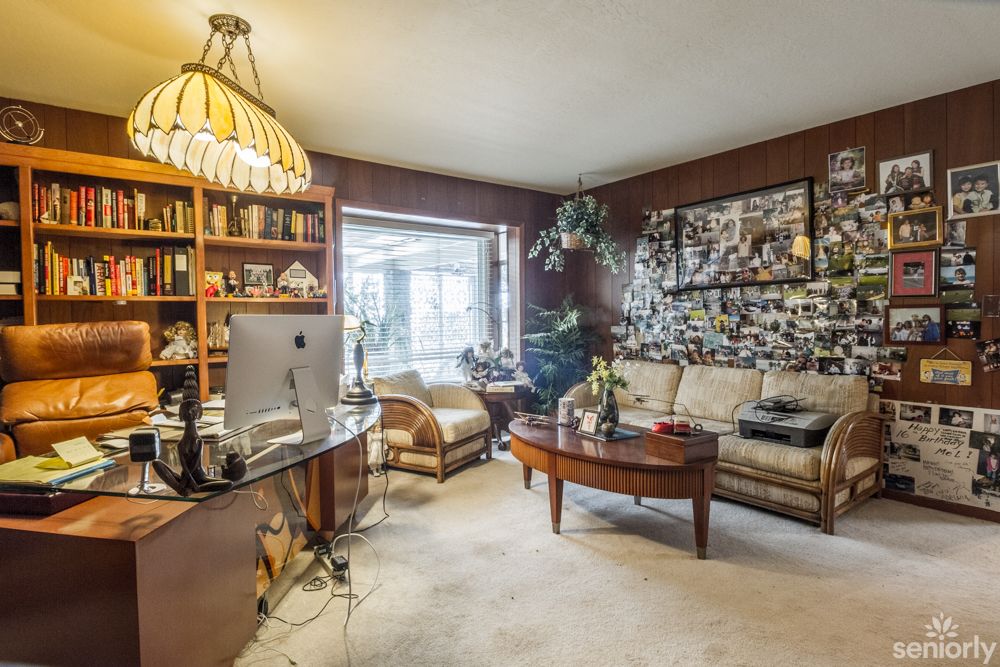 A cozy living room with a home office setup featuring a computer desk, sofa, shelves filled with books, and a large window allowing natural light to brighten the space.