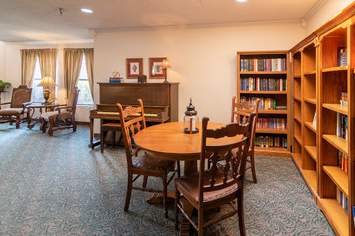 A cozy living room featuring a wooden dining table, chairs, a bookshelf filled with books, and a piano.