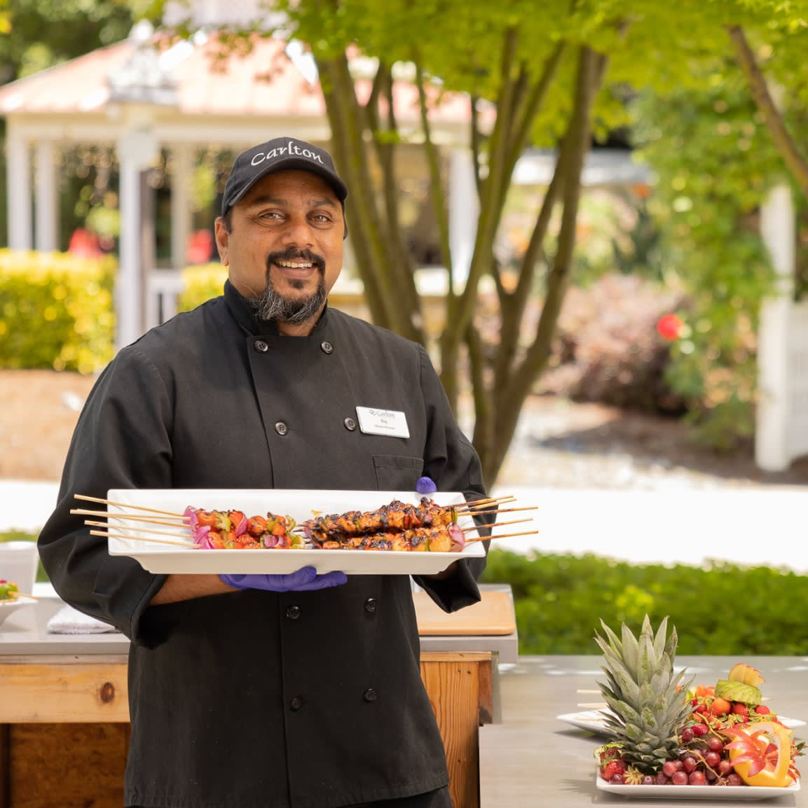A chef holds a platter of grilled skewers in an outdoor setting.