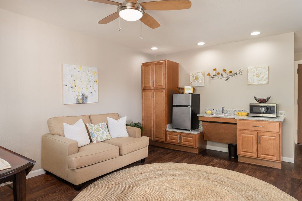 A cozy living room with a beige sofa, ceiling fan, small fridge, and wooden furniture.