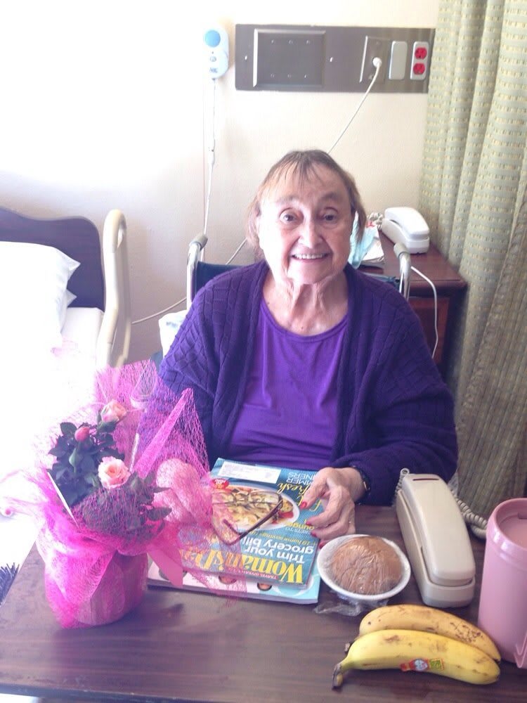 An elderly woman smiles while sitting at a table with flowers and a meal tray.