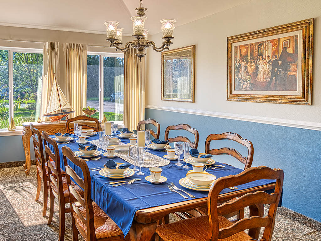 A well-appointed dining room featuring a wooden table set with a blue tablecloth, elegant dishware, and surrounded by matching chairs. The room is illuminated by a chandelier and decorated with framed artwork. Natural light pours in through the large wind